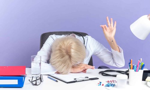 tired adult slavic female doctor in medical robe with stethoscope sitting at desk with office tools putting head on desk and gesturing ok sign isolated on purple background with copy space